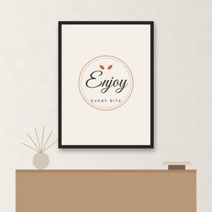 May include: A rectangular wall art print in a black frame featuring a white background with a centered circular design. The text "Enjoy" in a black script font sits above the text "EVERY BITE" in a smaller sans-serif font, with two small orange leaves above the word "Enjoy".