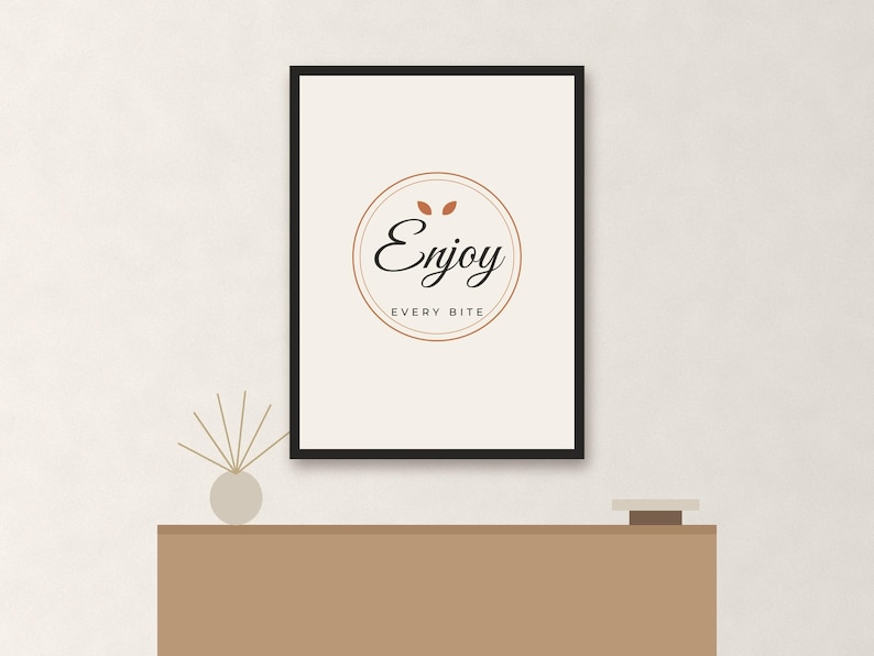 May include: A rectangular wall art print in a black frame featuring a white background with a centered circular design. The text "Enjoy" in a black script font sits above the text "EVERY BITE" in a smaller sans-serif font, with two small orange leaves above the word "Enjoy".