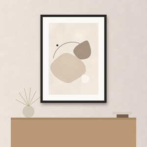 May include: A framed wall art print featuring an abstract composition of organic shapes in shades of beige, taupe, and white, accented by a thin black curved line and a small black dot, all enclosed within a simple black rectangular frame.