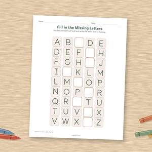 May include: A white educational worksheet titled Fill in the Missing Letters with a grid of alphabet letters in black ink. The page features empty boxes for students to complete the sequence. Several colored crayons are scattered around the paper.