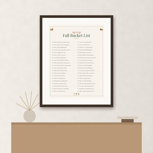 May include: A framed print titled My Cozy Fall Bucket List featuring a checklist of seasonal activities in dark text on a cream background. The design includes small autumn-themed illustrations at the top and bottom, enclosed in a dark brown frame.