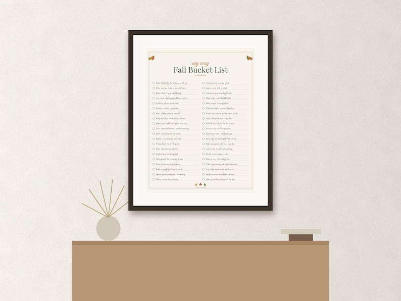 May include: A framed print titled My Cozy Fall Bucket List featuring a checklist of seasonal activities in dark text on a cream background. The design includes small autumn-themed illustrations at the top and bottom, enclosed in a dark brown frame.