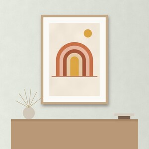 May include: A framed wall art print featuring a graphic illustration of a multi-colored rainbow arch in shades of terracotta, pink, and yellow, with a solid yellow circle representing the sun, all set against a light cream background.