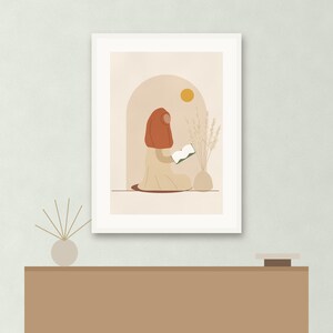 May include: A framed wall art print featuring a stylized illustration of a person wearing a rust-colored head covering and a beige garment, sitting and reading a white book. The scene includes a beige archway, a yellow sun, and a vase with dried grass.