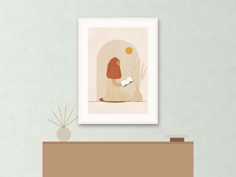 May include: A framed wall art print featuring a stylized illustration of a person wearing a rust-colored head covering and a beige garment, sitting and reading a white book. The scene includes a beige archway, a yellow sun, and a vase with dried grass.