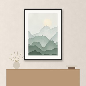 May include: A framed wall art print featuring a layered mountain landscape in shades of sage green and soft gray, with a pale yellow sun glowing in the sky. The artwork is displayed in a thin black frame, positioned above a solid brown console table.