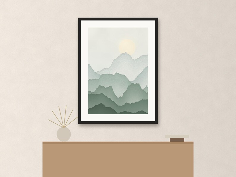 May include: A framed wall art print featuring a layered mountain landscape in shades of sage green and soft gray, with a pale yellow sun glowing in the sky. The artwork is displayed in a thin black frame, positioned above a solid brown console table.