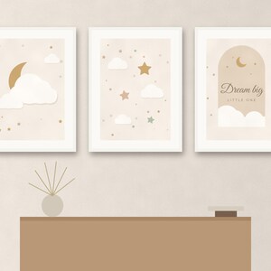 May include: Three framed wall art prints featuring a celestial theme with white clouds, gold crescent moons, and stars on a beige background. The third print includes the text Dream big LITTLE ONE inside an archway graphic design.