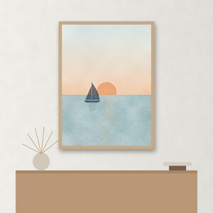 May include: A framed vertical wall art print featuring a dark blue sailboat on a textured light blue sea under a soft peach sky with a round orange sun. The artwork is displayed in a light wood frame, capturing a serene nautical scene.