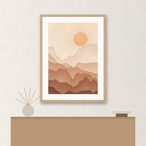 May include: A framed wall art print featuring a layered mountain landscape in shades of beige, tan, and terracotta. The artwork displays a circular sun positioned above the peaks, set within a light wood frame with a white mat border.