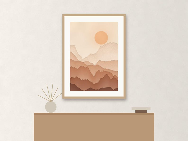 May include: A framed wall art print featuring a layered mountain landscape in shades of beige, tan, and terracotta. The artwork displays a circular sun positioned above the peaks, set within a light wood frame with a white mat border.
