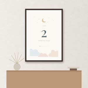 May include: A framed vertical poster with a cream background featuring a gold crescent moon and stars at the top. The text reads I AM 2 MONTHS OLD above a blank line for a date. The bottom displays light blue, pink, and beige cloud illustrations.