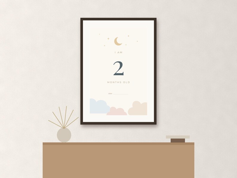 May include: A framed vertical poster with a cream background featuring a gold crescent moon and stars at the top. The text reads I AM 2 MONTHS OLD above a blank line for a date. The bottom displays light blue, pink, and beige cloud illustrations.