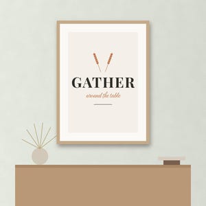 May include: A framed wall art print with a light beige background featuring two small orange wheat stalks above the black text GATHER and the smaller orange text around the table, with a horizontal line underneath the text.