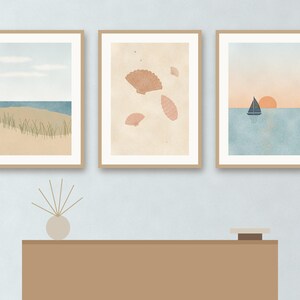 May include: Three framed wall art prints featuring coastal scenes. The left print shows a beach with dunes and grass. The center print displays various seashells on a beige background. The right print depicts a sailboat on water at sunset.