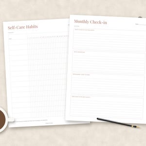 May include: Two white paper sheets featuring minimalist layouts. The left sheet is titled Self-Care Habits with a grid for tracking. The right sheet is titled Monthly Check-in with lined sections for reflection. A coffee cup and pen are present.
