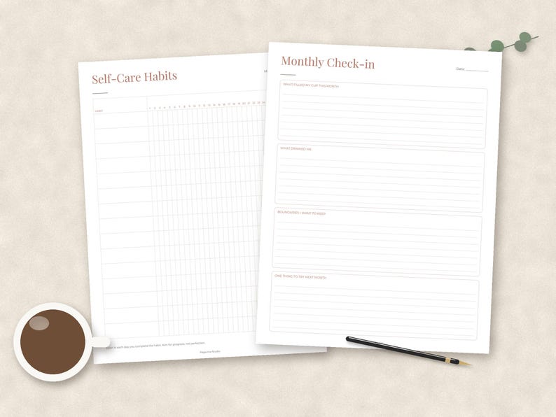 May include: Two white paper sheets featuring minimalist layouts. The left sheet is titled Self-Care Habits with a grid for tracking. The right sheet is titled Monthly Check-in with lined sections for reflection. A coffee cup and pen are present.