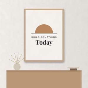 May include: A rectangular wall art print in a light brown frame featuring a white background with a tan semi-circle graphic above the black text "BUILD SOMETHING Today". The print is displayed above a solid light brown horizontal surface.