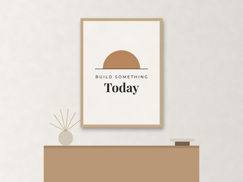 May include: A rectangular wall art print in a light brown frame featuring a white background with a tan semi-circle graphic above the black text "BUILD SOMETHING Today". The print is displayed above a solid light brown horizontal surface.