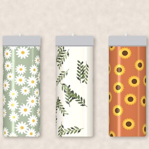 May include: Three cylindrical tumblers with flat gray lids. The left tumbler features a sage green background with white daisy patterns. The center tumbler has a cream background with green olive branch motifs. The right tumbler displays sunflowers.
