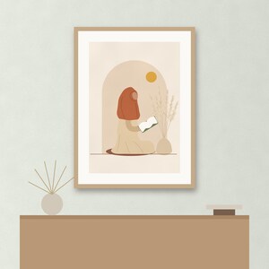 May include: A framed wall art print featuring a stylized illustration of a person wearing a rust-colored head covering and a beige garment, sitting and reading a book. The scene includes a yellow sun, an arched doorway, and a vase with dried grass.