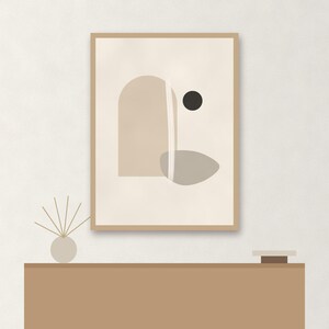 May include: A framed wall art print featuring an abstract composition with a beige arch, a gray organic shape, a small black circle, and a vertical white line on a cream background, displayed above a brown console table with a reed diffuser.