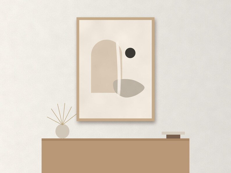 May include: A framed wall art print featuring an abstract composition with a beige arch, a gray organic shape, a small black circle, and a vertical white line on a cream background, displayed above a brown console table with a reed diffuser.