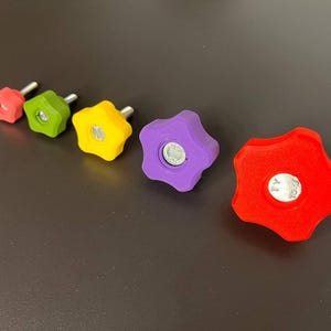 Peut inclure: Un ensemble de cinq boutons en forme d'&eacute;toile de tailles gradu&eacute;es, comprenant un rouge, un vert, un jaune, un violet et un plus grand rouge. Chaque bouton poss&egrave;de un boulon m&eacute;tallique central avec une tige filet&eacute;e d&eacute;passant &agrave; l'arri&egrave;re.