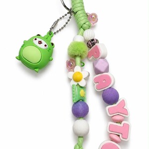 Puede incluir: Un llavero de paracord verde lima con un dije de rana verde colgante, una cuenta de flor blanca y varias cuentas texturizadas en morado, blanco y verde. El llavero incluye cuentas de letras rosas que deletrean el nombre RAYTO, unido a una argolla.