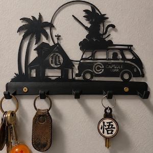 May include: A black metal wall-mounted key holder featuring a silhouette of a palm tree, a building with a Japanese character, a van labeled Capsule Corp, and a character on a cloud. It has five hooks, two gold screws, and holds three keychains.
