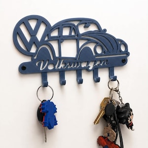 Puede incluir: Un soporte para llaves de pared en color azul mate que presenta una silueta recortada de un coche Volkswagen Beetle clásico y el logotipo de Volkswagen. El diseño incluye la palabra Volkswagen en cursiva y cinco ganchos con dos llaveros.
