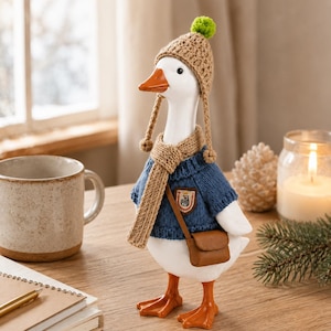 May include: A white goose figurine wearing a tan crocheted hat with a green pom-pom, a matching tan scarf, and a blue knitted sweater. The goose has an orange beak and feet, and carries a small brown crossbody bag with a crest patch on the chest.