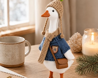 May include: A white goose figurine wearing a tan crocheted hat with a green pom-pom, a matching tan scarf, and a blue knitted jumper. The goose has an orange beak and feet, and carries a small brown crossbody bag with a crest patch on the chest.
