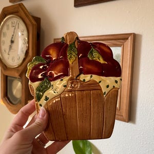 May include: A ceramic wall-mounted basket holder featuring a brown wood-grain texture, filled with glossy red apples and green leaves. The basket is draped with a cream-colored cloth patterned with small green dots, creating a decorative kitchen accent.