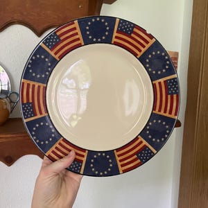 May include: A round ceramic plate with a cream-colored center and a wide decorative border. The border features an alternating pattern of rectangular American flag motifs and dark blue circular sections adorned with a ring of white stars.