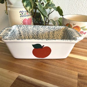 May include: White ceramic loaf dish with a red apple design and green leaf. The rim has a blue and grey speckled pattern. A decorative mug with fruit and floral designs is in the background.