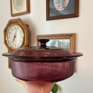May include: A deep purple glass casserole dish with a matching lid. The dish features a vertical ribbed texture along the exterior sides and a smooth, flat rim. The lid has a central knob handle and a slightly domed shape with a smooth surface.