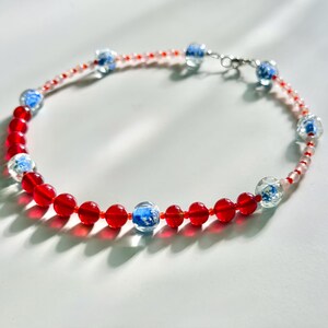 May include: A beaded bracelet with alternating red and blue glass beads. The bracelet has a delicate design with small red beads and larger, clear beads with blue accents. The clasp is silver-toned.