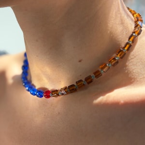 May include: A beaded necklace with a colour gradient. The necklace transitions from blue beads to red and then to amber-coloured square beads. The necklace is worn around the neck.