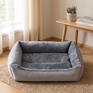 May include: A rectangular gray pet bed with a plush, faux fur interior and a soft, suede-like exterior. The bed is designed with raised sides for added comfort and security. It sits on a woven, natural fiber rug.