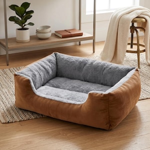 Peut inclure: Un lit pour animaux de compagnie rectangulaire avec un extérieur marron et un intérieur en peluche grise. Le lit a un profil bas et est conçu pour le confort. Le lit est posé sur un sol en bois avec un tapis en dessous.