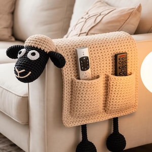 Peut inclure: Un organiseur de bras de canapé en crochet beige en forme de mouton avec deux poches. Le mouton a un visage, des oreilles et des pattes noirs, et une tête beige. Deux télécommandes sont dans les poches.