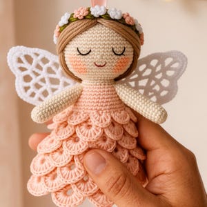 Peut inclure: Une poupée fée au crochet faite à la main, avec une robe pêche et des ailes en dentelle blanche. La poupée a une couronne florale et un ruban pour la suspendre. La poupée est tenue dans une main.
