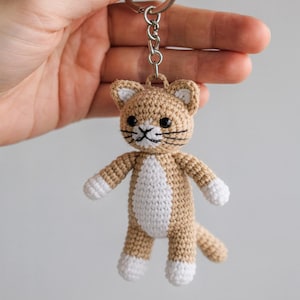 Peut inclure: Porte-clés chat crocheté beige et blanc. Le chat fait main a des yeux noirs, des moustaches et une poitrine et des pattes blanches. Le porte-clés est fixé à un anneau en métal argenté.