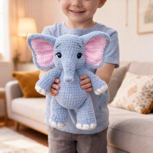 Peut inclure: Une peluche éléphant en crochet bleu clair avec des oreilles intérieures roses et des détails blancs sur les pieds et la trompe. L'éléphant a des yeux noirs et est tenu par une personne.