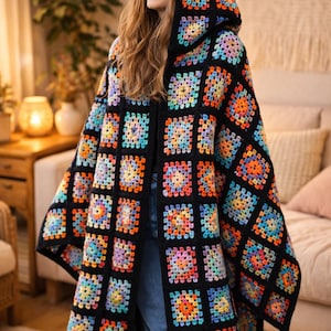Peut inclure: Un poncho à capuche en crochet à devant ouvert, de conception patchwork. Le poncho présente une base noire avec des carrés colorés dans les tons orange, jaune, bleu et violet. Le vêtement est porté par une personne dans un cadre domestique.