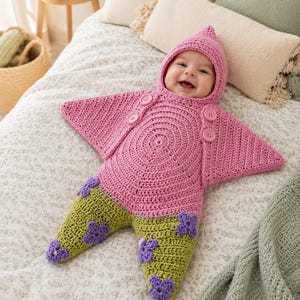 May include: A pink and green crocheted star-shaped baby bunting with a hood. The upper section is pink with four matching buttons, while the lower section is green with purple crocheted flower appliqués attached to the legs and pointed feet.