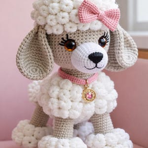 Peut inclure: Un chien caniche au crochet avec un corps blanc, des oreilles grises et un nœud rose. Le chien porte un collier rose avec un pendentif doré. Le caniche est assis sur une surface rose. La fourrure est faite de petits points de crochet texturés.
