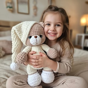 Peut inclure: Un ours en peluche crocheté à la main, de couleur crème, beige et blanc. L'ours porte un long bonnet de nuit crème et a des pattes et des oreilles beiges. Le jouet en peluche est tenu par un enfant souriant.