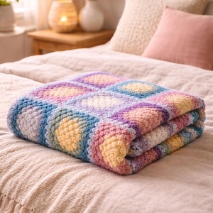 Può includere: Coperta all'uncinetto piegata, colorata. La coperta presenta un motivo di cerchi nelle tonalità del giallo, rosa, viola, blu e bianco. È piegata ordinatamente su un letto con un copriletto e cuscini di colore neutro.